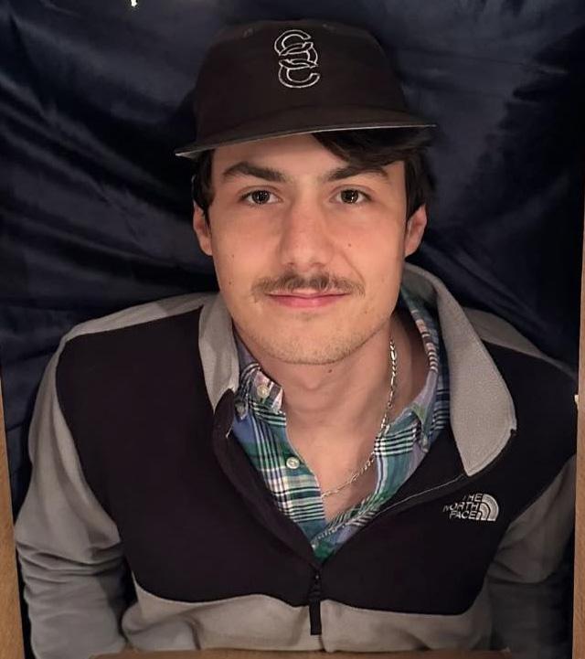 Young man in a dark North Face jacket and cap sits smiling, with a blue plaid shirt underneath