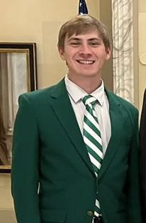 Young man in a green blazer and striped tie smiling in what appears to be a government building.