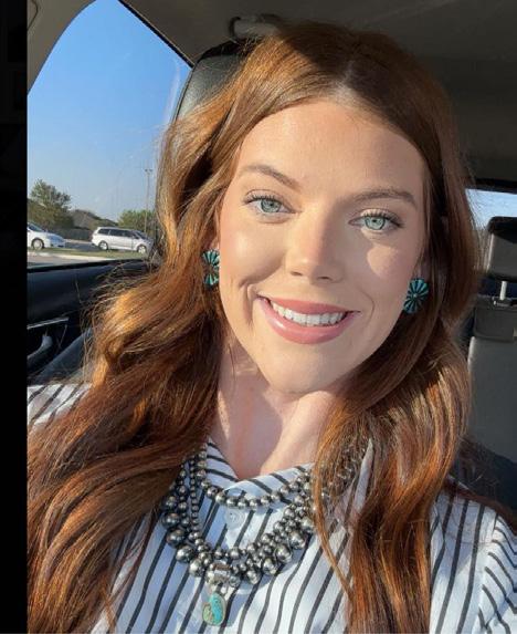 Young woman with long auburn hair sitting in a car, smiling, wearing turquoise earrings and a striped shirt.