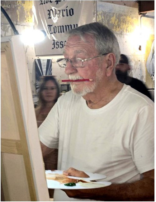 Elderly artist Robert Wright painting during a live competition, holding a palette and using a paintbrush in his mouth while focusing on his canvas.