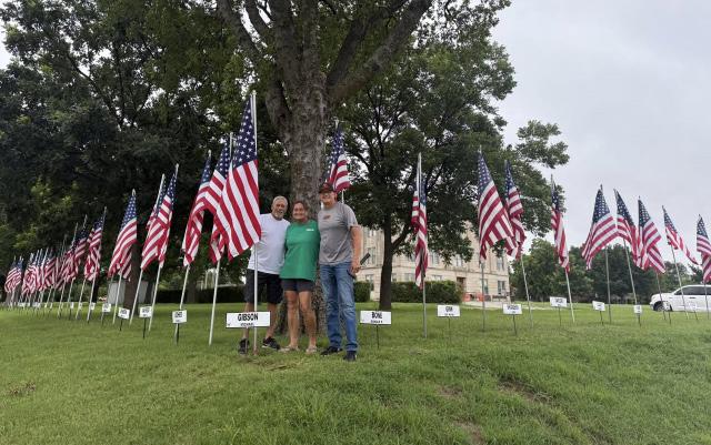 Three Rotary Club members pose together beneath a large tree, surrounded by American flags and name markers during a community Fourth of July celebration.