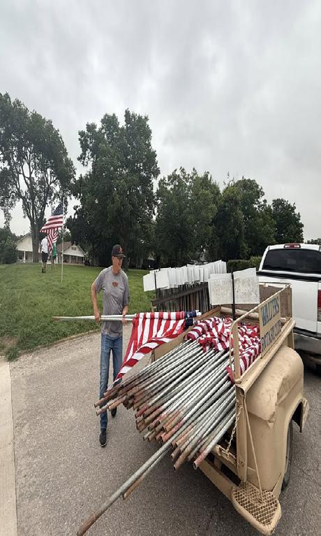 A volunteer loads flagpoles into a trailer marked “WALTERS ROTARY CLUB” as flags begin to go up along a neighborhood street.