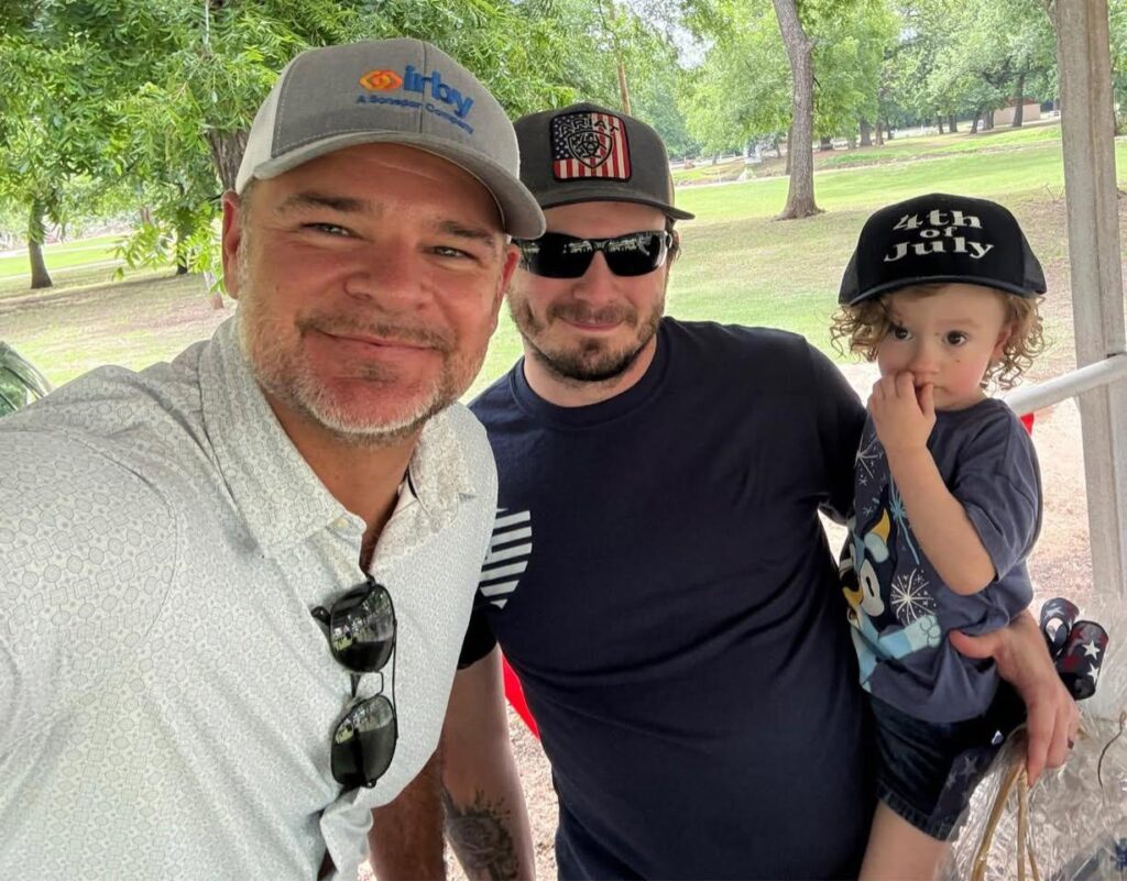 Three generations smile for a photo: a man in an Irby cap, another adult in a “4th of July” hat, and a toddler wearing patriotic gear, all celebrating the holiday together.