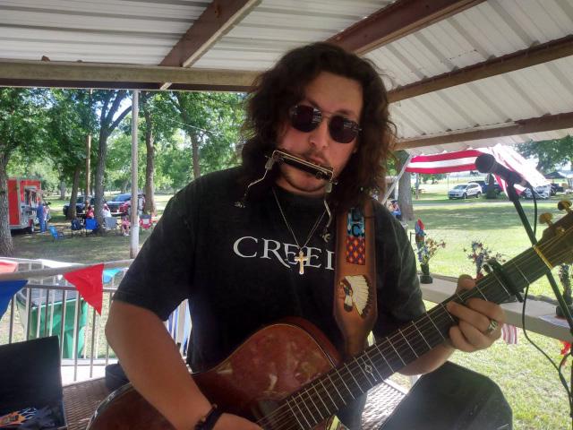 A musician wearing sunglasses plays guitar and harmonica on stage, performing live under a covered pavilion decorated for Independence Day.