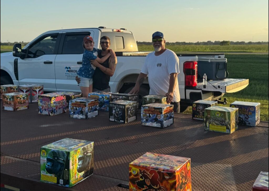 Three people pose behind a flatbed trailer loaded with large fireworks boxes, preparing for the community fireworks show at sunset, with a white pickup truck and open field in the background.