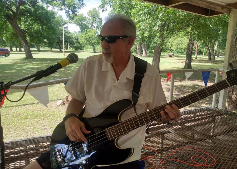 A musician in sunglasses plays an electric bass guitar and sings into a microphone from a shaded outdoor stage at the park celebration.