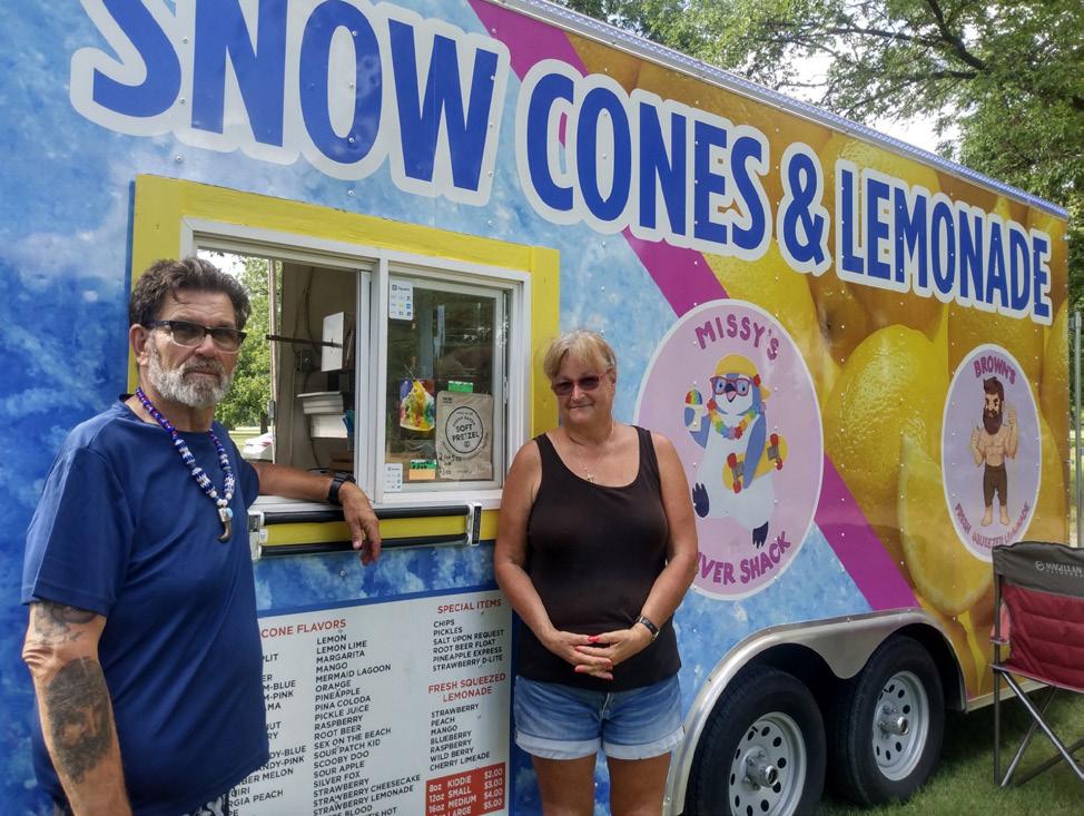 A man and woman stand in front of a colorful “Snow Cones & Lemonade” food truck, decorated with cartoon mascots and a vibrant menu.