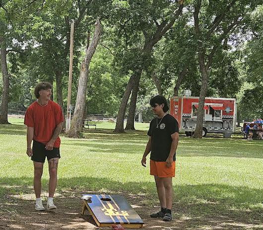 Two teens play a game of cornhole in a shaded area of Sultan Park, with a red food truck in the background.