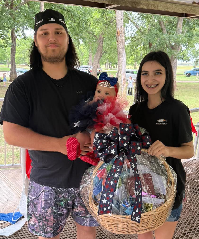A young couple poses with a baby dressed in a red, white, and blue tutu and tiara, holding a patriotic gift basket wrapped in cellophane with a large star-spangled bow.
