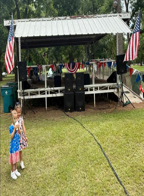 Two young girls stand in front of a stage decorated with American flags and bunting, holding flags and dressed in festive outfits during the 4th of July celebration.