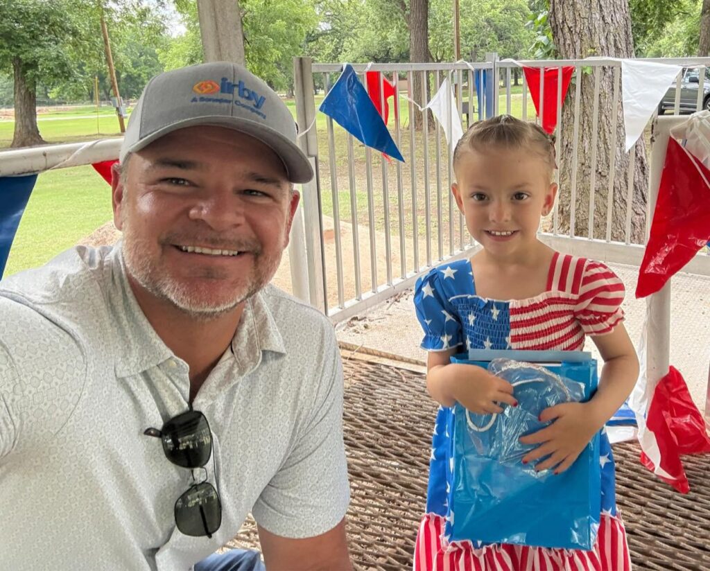 A smiling man in a ballcap takes a photo with a little girl dressed in a red, white, and blue dress holding a blue prize bag, surrounded by patriotic bunting.