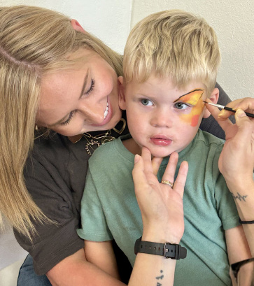 A blonde woman smiles while holding a young boy whose face is being painted with a colorful butterfly during an elementary school event.