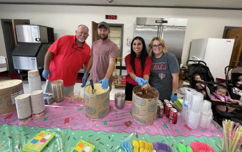 Four adults stand behind a colorful ice cream station with large tubs of ice cream, toppings, and cones at a school event. A woman in the background tends to young children in a stroller. The table is covered with a bright pink and green sprinkle-themed tablecloth.