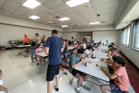 Four adults stand behind a colorful ice cream station with large tubs of ice cream, toppings, and cones at a school event. A woman in the background tends to young children in a stroller. The table is covered with a bright pink and green sprinkle-themed tablecloth.