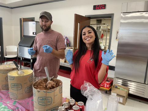 Four adults stand behind a colorful ice cream station with large tubs of ice cream, toppings, and cones at a school event. A woman in the background tends to young children in a stroller. The table is covered with a bright pink and green sprinkle-themed tablecloth.